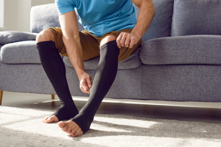 Man putting on black compression stockings in a living room setting, demonstrating the use of graduated compression socks for enhancing leg circulation and preventing health issues like varicose veins, shown as part of a routine for promoting leg health.